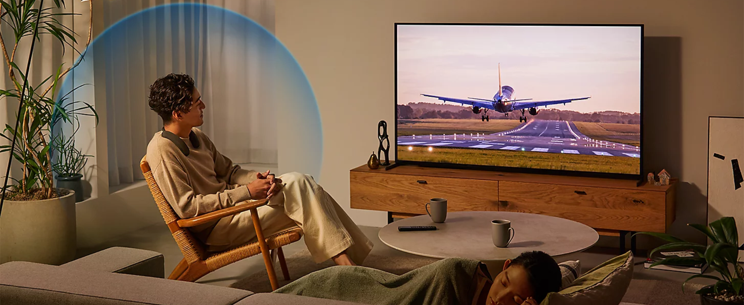 Person sitting on a chair in a living room wearing BRAVIA Theatre U while watching a movie on a big screen TV with screenshot of an airplane landing and person sleeping on sofa in foreground