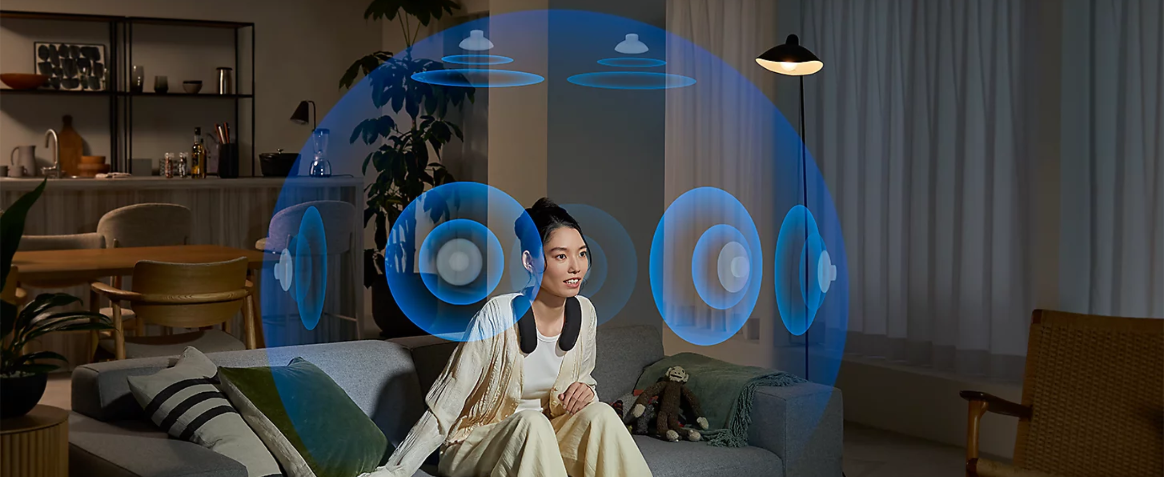 Person sitting on a grey sofa in a living room wearing BRAVIA Theatre U while watching a movie with a blue sphere of phantom speakers all around