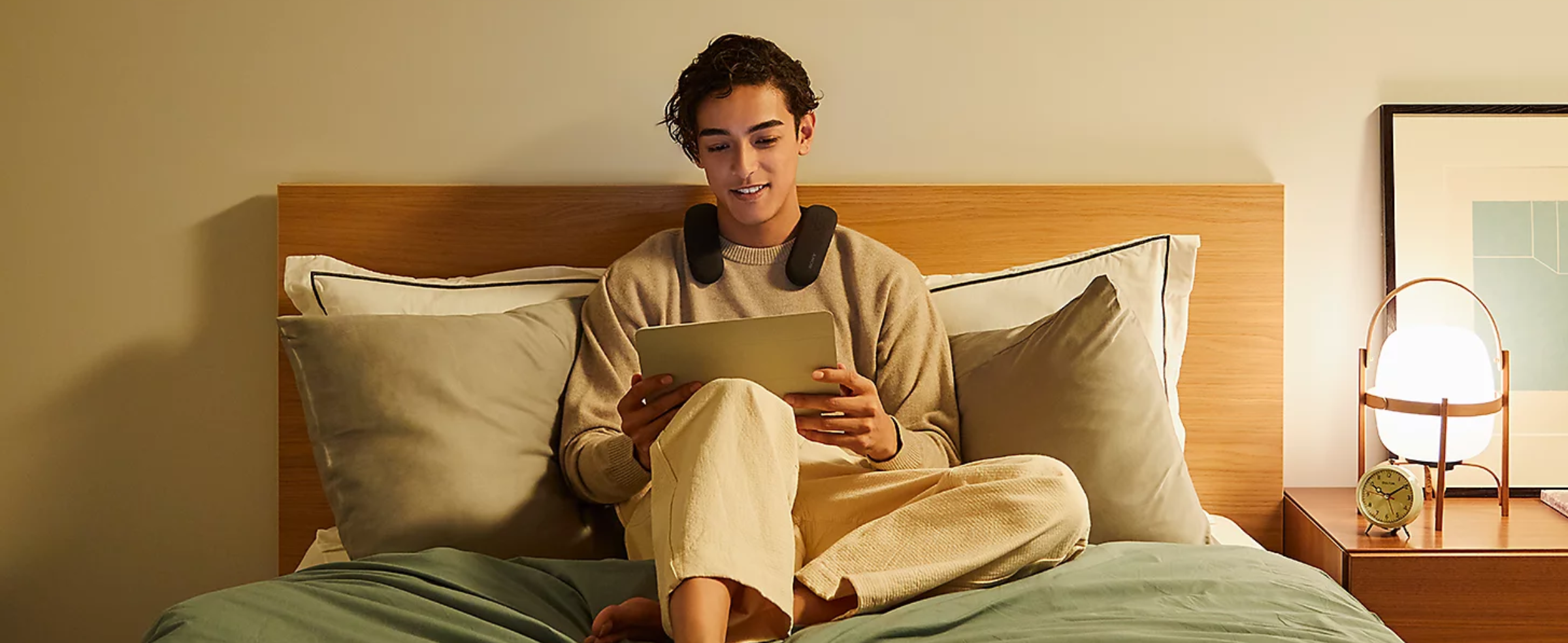 Person sitting on a bed wearing BRAVIA Theatre U while watching content on a tablet with a bedside table to right with lamp and clock