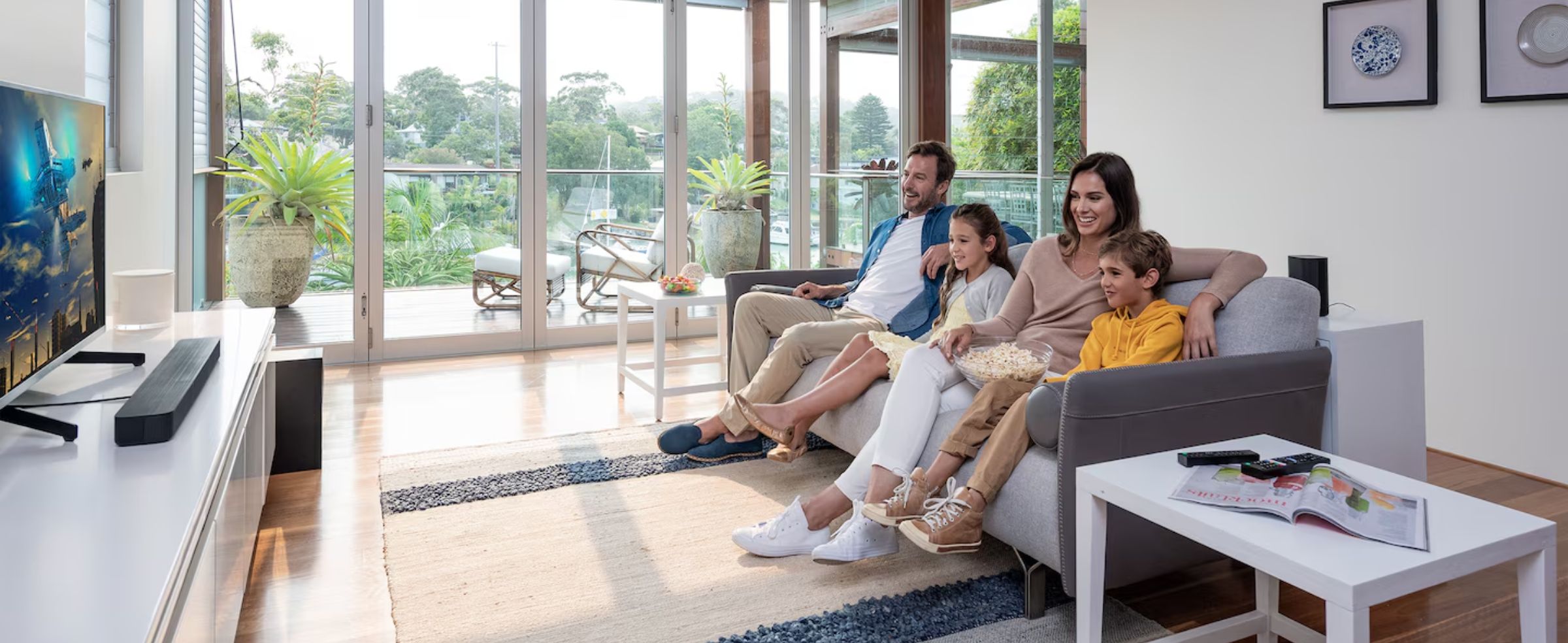 A family of four is sitting on a couch, watching TV, with a soundbar and sub-woofer.